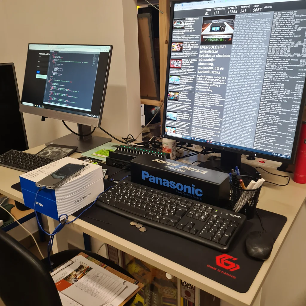 Két nagy monitor, billentyűzet, egér, Panasonic feliratú egérpad, dobozok és irodai eszközök egy asztalon.